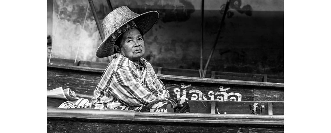 A vendor wearing her traditional mo hom apparel and ngob hat offers goods for sale from her sampon at Damnoen Saduak 