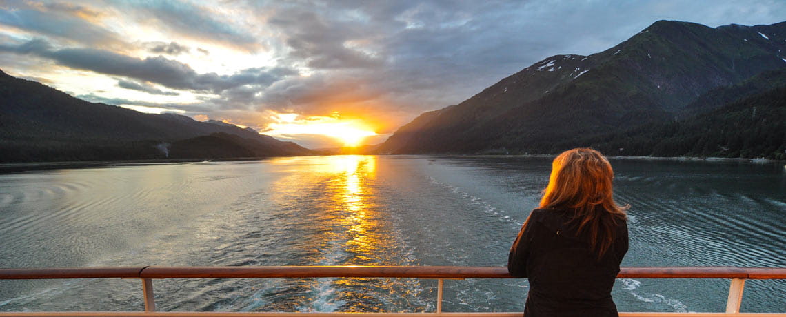 Woman staring at sunset while standing near railing on back of cruise ship