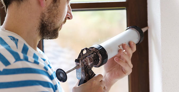 A young man in a striped shirt seals out window leaks and drafts with a caulk gun.