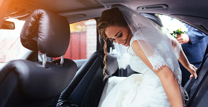 Followed by her groom, a brunette bride slides into the back seat of a town car. 