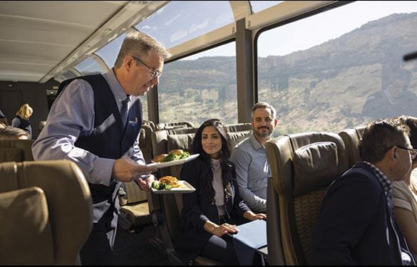 Tourists aboard a train with large windows for a scenic view, receiving a meal from their server