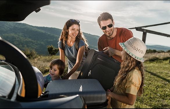 Family with two adults and two kids loading suitcases into back of vehicle, with mountains and sun in the distance.