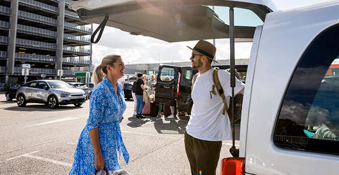 Mother and son packing up a rental van in France where they are vacationing to. They are standing in a car park at an airport in the sun.