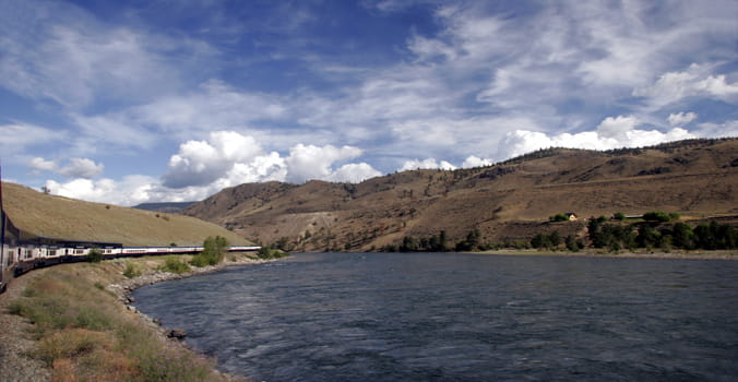 Panoramic view of a Rocky Mountaineer train passing a river and mountains