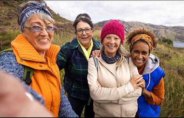 Front view of a small group of mature and senior female friends hiking up the mountain landscape surrounding Loch Torridon in the Northwest Highlands, Scotland. A senior woman is wearing a wooly hat and the rest are wearing bandanas, they are side by side taking a selfie on a smartphone.