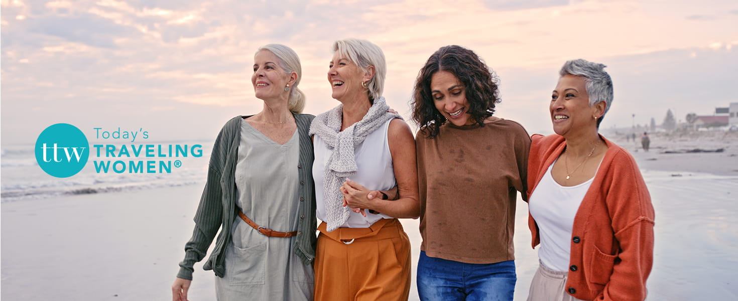 Group of women friends together smiling and standing on the beach. Today's Traveling Women logo.
