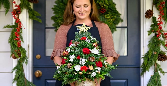 A woman standing in front of a festive house door holding a Christmas tree floral arrangement
