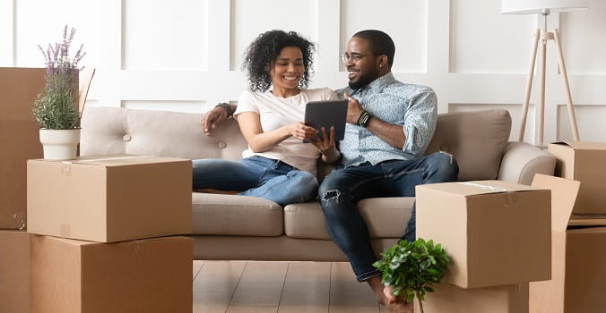 A woman and man sitting on a couch looking at a tablet with moving boxes all around 