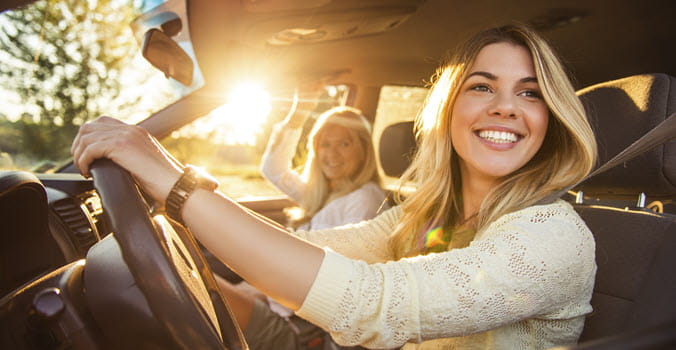Young woman with blond hair smiling while behind the wheel of a car with her young woman friend with blond hair in the passenger seat. A sunset appears in the background.