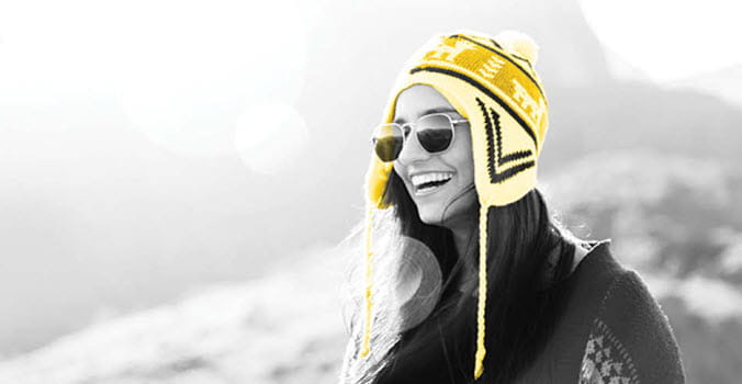 Black and white photo of a woman wearing sunglasses and a yellow winter hat with a big smile on her face