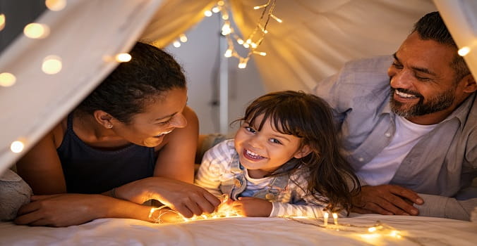 Little girl talking to mother and father lying on bed in illuminated tent made of sheets.