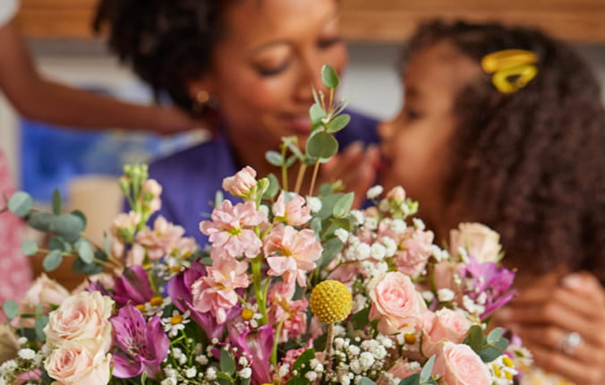 A mom hugs her daughter, with beautiful Mother's Day flowers in the foreground