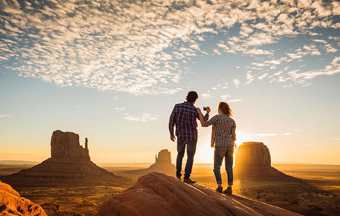 Two people stand on a rock at sunset, holding hands, with large rock formations in the background under a partly cloudy sky.