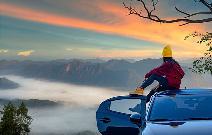 Young woman sits on top of vehicle overlooking a scenic mountain view