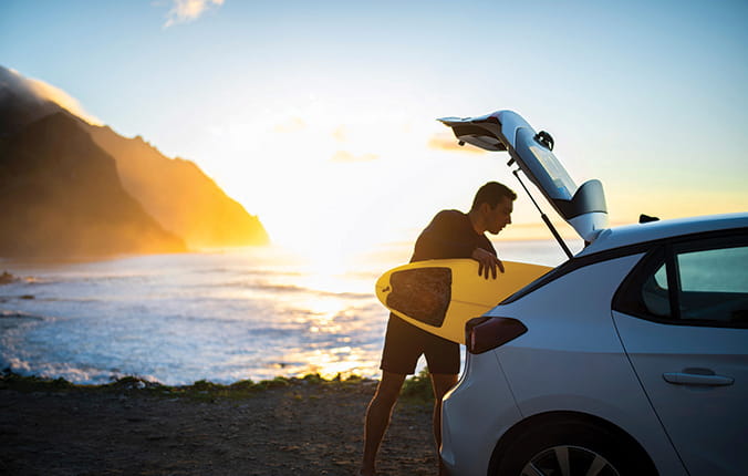 Man takes his yellow surfboard out of his car, with sunrise over the water in background