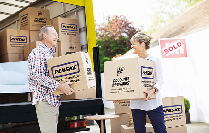 A man and woman carry boxes labeled 'Penske' from a moving truck. More boxes are inside the truck and scattered nearby. A 'Sold' sign is visible in the background.