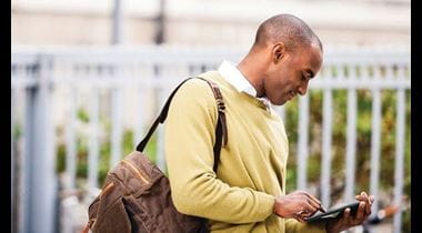 Man standing outside wearing a messenger bag looking down at his tablet 