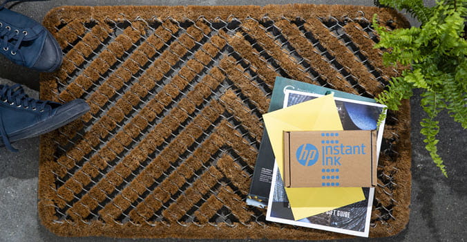 Front door mat with HP instant ink package on the doorstep with a plant to the right and a pair of shoes to the left