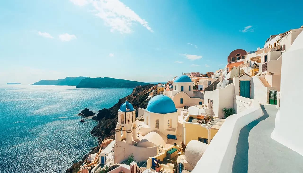 A sunlit view of the iconic whitewashed buildings and blue-domed churches of Santorini, Greece, overlooking the deep blue sea.