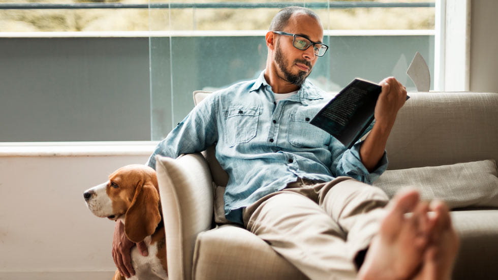 Man sitting on a couch with his feet up, reading a book, and petting a dog