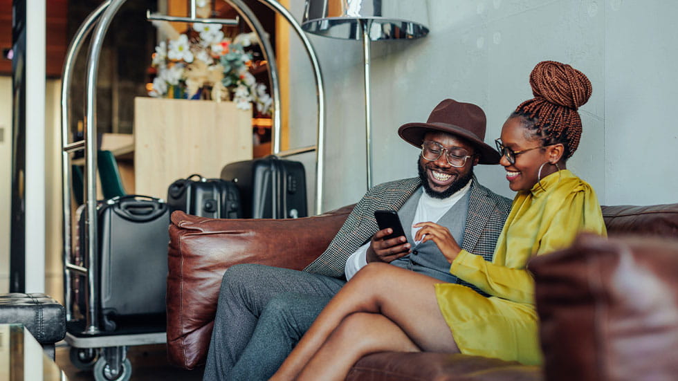 A wealthy couple is lounging in the hotel as they're waiting to be taken to their luxurious suit in the hotel. They are sitting on a sofa with their luggage on the cart and are using a smartphone.