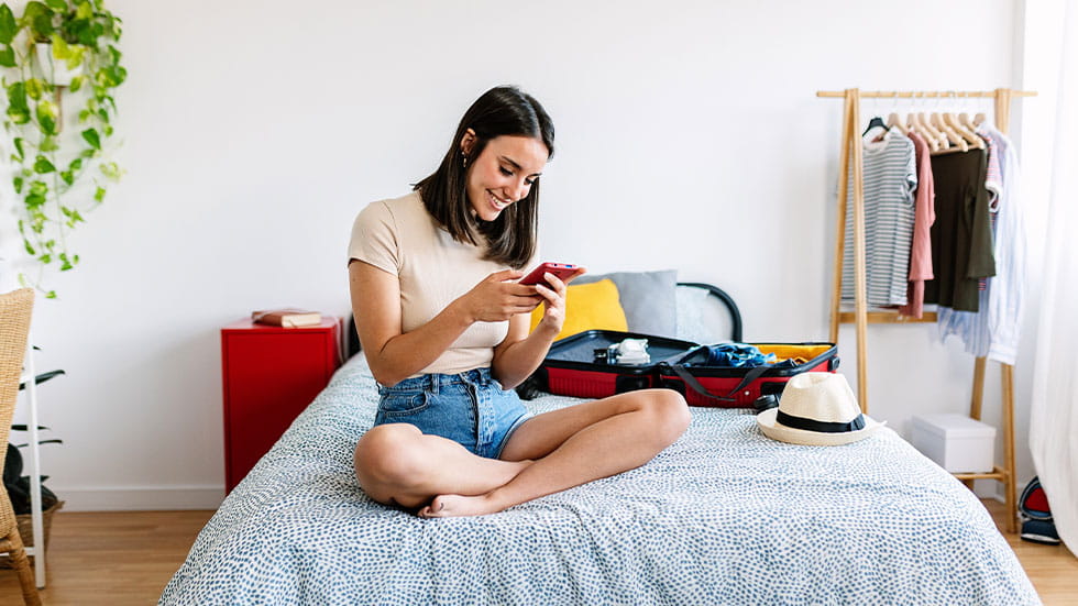 woman sitting on bed using phone next to suitcase