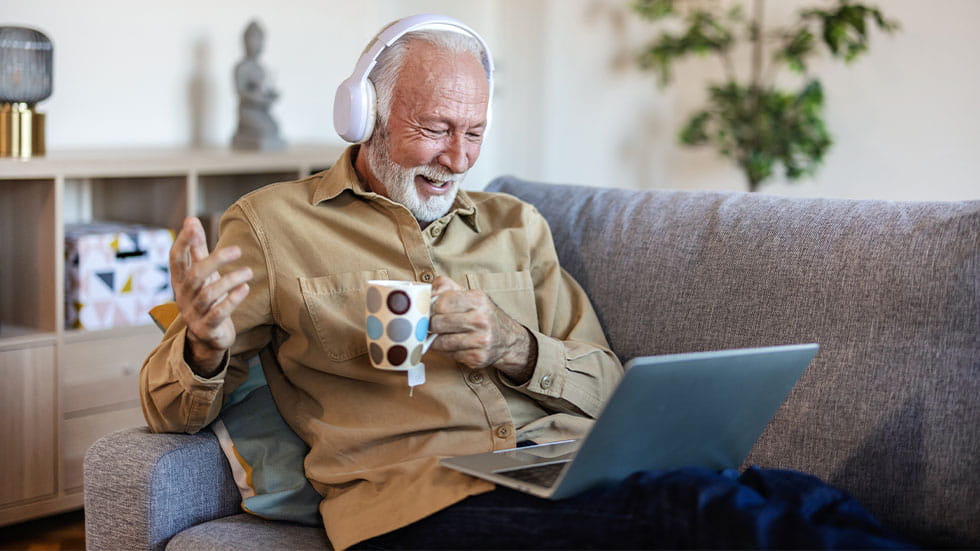 elder man wearing headphones and looking at laptop on couch