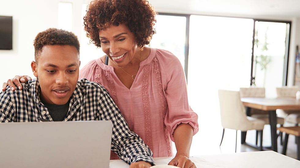 son looking at laptop with mother looking over his shoulder smiling